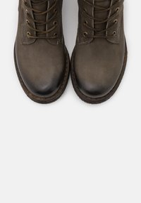 Botas de tobillo de cuero marrón con puntera redonda, suela plana y seis ojales metálicos decorativos para los cordones, con una textura suave y costuras sutiles.