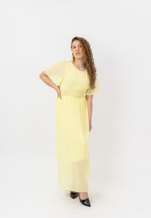 Mujer con el pelo rizado que lleva un vestido largo amarillo y zapatos negros puntiagudos, de pie con una mano en la cadera sobre un fondo blanco.