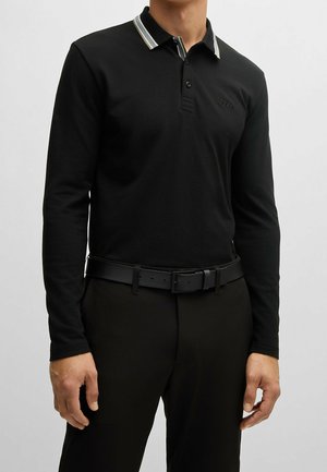 Mann trägt ein schwarzes Langarm-Poloshirt mit kontrastierendem Kragen und schwarzen Gürtel mit schwarzer Hose.