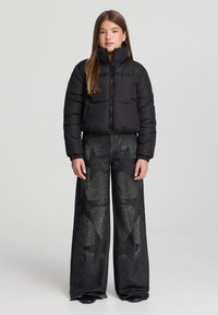 Giacca a vento nera con colletto alto e chiusura con zip, abbinata a jeans neri a gamba larga con motivi a stelle e dettagli borchiati.