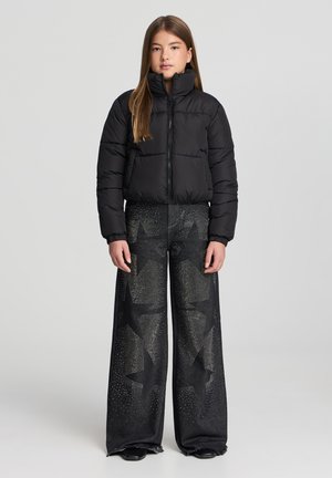 Giacca a vento nera con colletto alto e chiusura con zip, abbinata a jeans neri a gamba larga con motivi a stelle e dettagli borchiati.