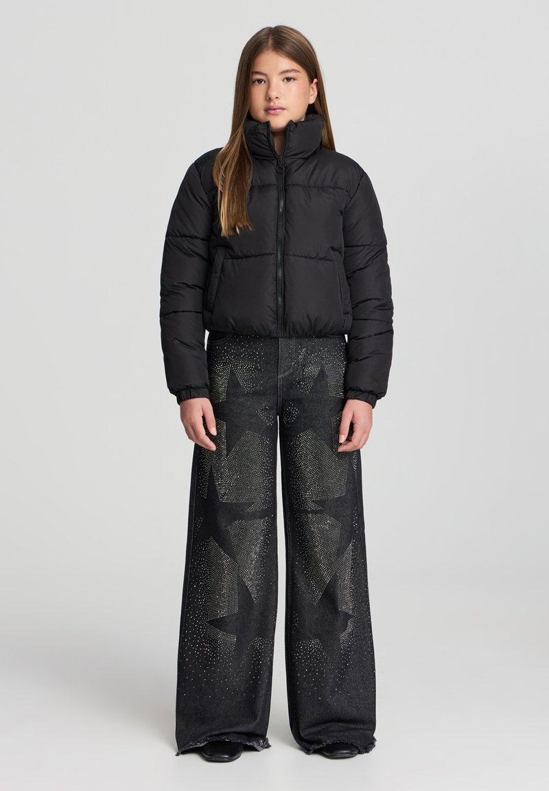 Giacca a vento nera con colletto alto e chiusura con zip, abbinata a jeans neri a gamba larga con motivi a stelle e dettagli borchiati.