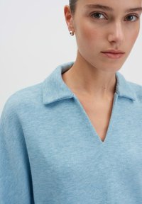 Vrouw in een lichtblauwe gebreide top met een V-hals en kleine zilveren ringoorbellen, weergegeven van de nek omhoog.