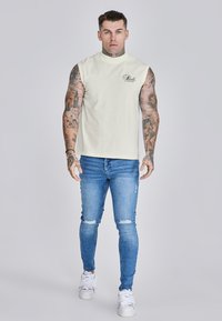 Camiseta sin mangas blanca con cuello alto y logo negro, combinada con jeans skinny desgastados azules y zapatillas blancas.