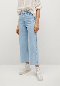 Personne portant un jean large et court bleu clair, des baskets montantes blanches, une chemise crème à boutons, et des bagues dorées à la main droite.