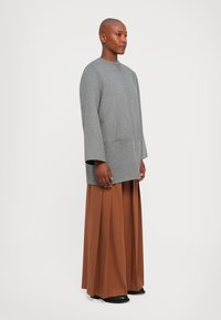 Persoană cu cap ras, îmbrăcată într-un pulover oversized gri și pantaloni maro cu picioare largi, stând în fața unui fundal alb simplu.