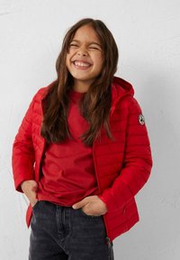 Rode gewatteerde jas met capuchon, textuurmatige doorgestikte pasvorm en logo-embleem, gedragen over een bijpassend rood shirt en donkergrijze spijkerbroek.