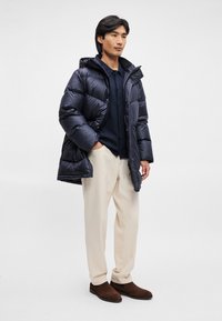 Casaco puffer azul-marinho com capuz, apresentando um acabamento brilhante e um design acolchoado, combinado com calças creme e botins castanhos.