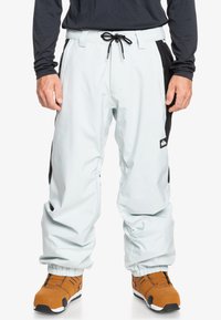 Pantalones de nieve azul claro con paneles laterales negros, cintura elástica con cordón y puños elásticos en los tobillos, combinados con botas de snowboard marrones.