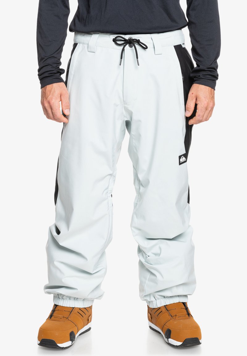 Pantalones de nieve azul claro con paneles laterales negros, cintura elástica con cordón y puños elásticos en los tobillos, combinados con botas de snowboard marrones.