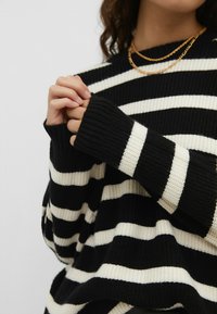 Personne portant un pull en tricot rayé noir et blanc, les mains jointes près de la poitrine et deux colliers en chaîne dorée autour du cou.