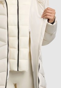 Biela prešívaná pufferová bunda s kapucňou, s úplným zipsom, kontrastnými čiernymi zipsami a hladkým vonkajším povrchom.