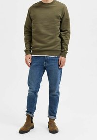 Olivově zelená mikina s kulatým výstřihem a žebrovanými manžetami; doplněná modrými džínami s ohrnutými lemy. Hnědé kotníkové boty s kulatou špičkou.