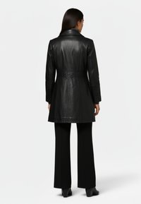 Kvinna som bär en figursydd svart läderkappa till knäna, svarta byxor och svarta klackskor, vänd bort från kameran mot en enfärgad vit bakgrund.