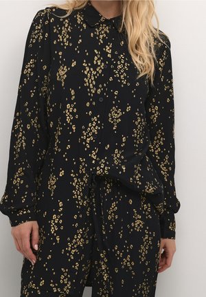 Femme portant une chemise noire à manches longues boutonnée et un pantalon avec de petits motifs floraux dorés, debout contre un mur blanc uni.