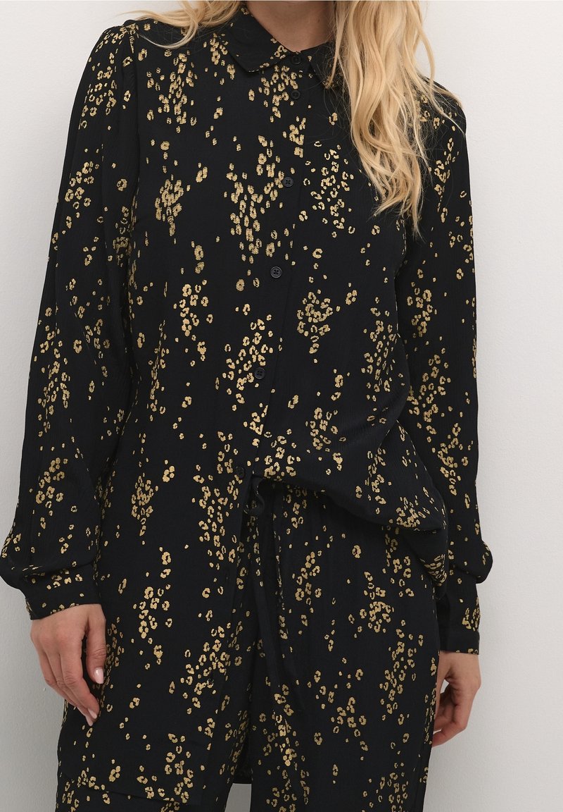 Femme portant une chemise noire à manches longues boutonnée et un pantalon avec de petits motifs floraux dorés, debout contre un mur blanc uni.