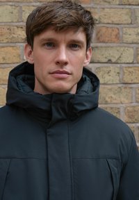 Veste noire rembourrée avec un col haut et une capuche. Présente une texture lisse et un design minimaliste avec des coutures avant. Adaptée aux temps froids.