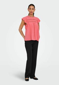Blusa rosa chiaro con maniche corte a volant e motivi testurizzati. Abbinata a pantaloni neri e scarpe basse. Design semplice e dal taglio morbido.