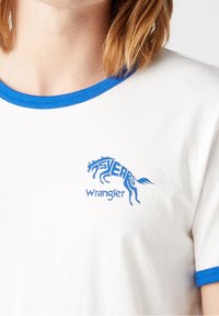 Vit t-shirt med blå rund halsringning och ärmslut. Har en blå grafik med en häst och texten "5 ÅR Wrangler" tryckt på bröstet.
