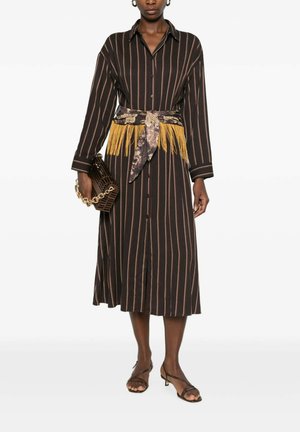 Donna che indossa un vestito marrone a righe con maniche lunghe e cintura floreale con frange gialle, che tiene una borsa marrone con tracolla a catena dorata.
