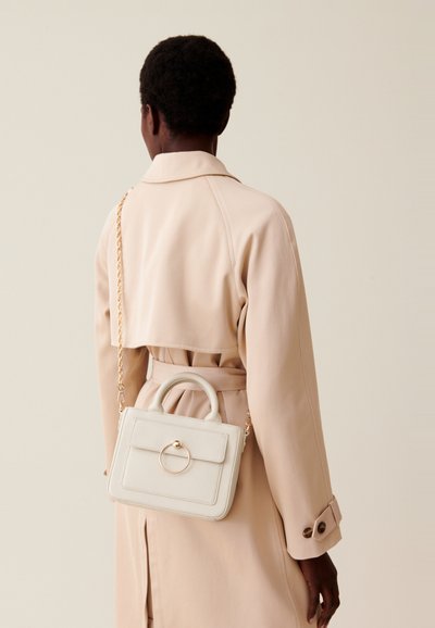 Sac à main en cuir blanc avec une forme structurée, une poignée supérieure et des accessoires ronds en or. Porté avec un trench-coat beige, la chaîne d'épaule est visible.