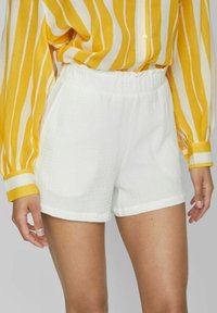 Bílé látkové šortky s elastickým pasem, se strukturovaným, lehkým designem a ohrnutým lemem. V kombinaci se žluto-pruhovanou blůzou.