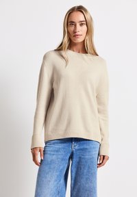 Beżowy sweter z dzianiny o okrągłym dekolcie, z opuszczonymi ramionami i prążkowanymi mankietami, zestawiony z jasnoniebieskimi spodniami typu wide-leg.