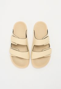 Beige-liukumäntävät sandaalit, joissa on kolme säädettävää tarranauhaa, sileä synteettinen materiaali, muotoiltu pohja ja hillityt mustat aksentit.
