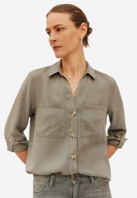 Lichtgrijze button-up shirt met twee borstzakken, opgerolde mouwen en een zachte textuur. Heeft parelachtige knopen en een gestructureerde kraag.