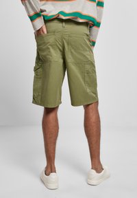 Man som bär olivgröna knälånga cargoshorts och vita sneakers, med en hand i bakfickan, stående mot en enfärgad ljusgrå bakgrund.
