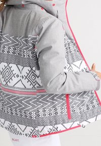 Graue Jacke mit einem gemusterten Rücken, der schwarz-weiße geometrische Designs zeigt, akzentuiert durch rosa Reißverschlüsse und Besätze. Mit Kapuze.