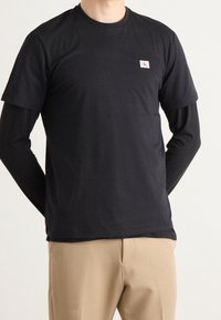 Czarna koszulka z krótkim rękawem noszona na czarnej bluzie z długim rękawem; ma małe białe i czerwone logo na piersi. Gładki materiał, luźny krój.