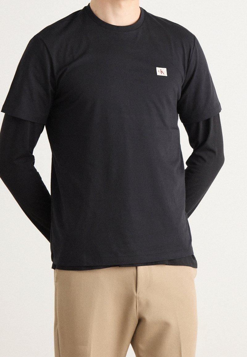 Czarna koszulka z krótkim rękawem noszona na czarnej bluzie z długim rękawem; ma małe białe i czerwone logo na piersi. Gładki materiał, luźny krój.