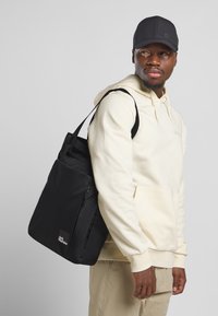 Zwarte schoudertas van duurzaam stof met verstelbare schouderbanden. Voorzien van een voorklep met rits en een logo-embleem. Gekoesterd over een licht hoodie.