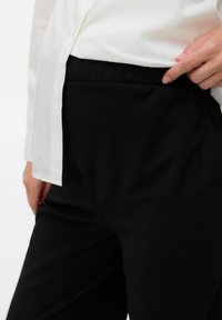 Pantalon noir en tissu lisse, taille haute, avec poches latérales, associé à une chemise blanche à boutons, mettant en valeur un design élégant.