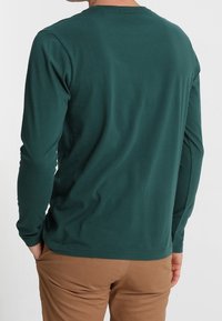 Mann in einem dunkelgrünen Langarmhemd und braunen Hosen, mit den Händen in den Taschen, von der Kamera wegstehend.