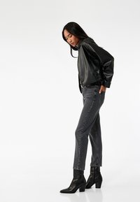 Giacca bomber nera in pelle con polsini elasticizzati, abbinata a jeans grigi a vita alta e stivaletti neri con tacco. Design semplice e moderno.