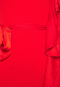 Robe rouge présentant une texture côtelée, une large ceinture en tissu et un détail de manche à volants. Le matériau semble lisse avec un léger éclat.