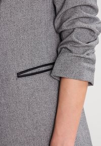 Szary teksturowany żakiet o dopasowanym kroju. Posiada czarną kieszeń wykończoną lamówką oraz marszczone rękawy, co dodaje głębi projektowi.