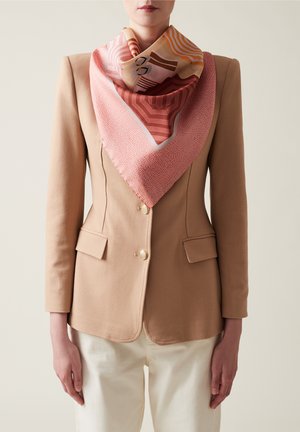 Foulard en soie dans des tons roses et oranges, arborant des motifs géométriques. Porté avec un blazer beige, mettant en valeur une texture lisse et une forme triangulaire.