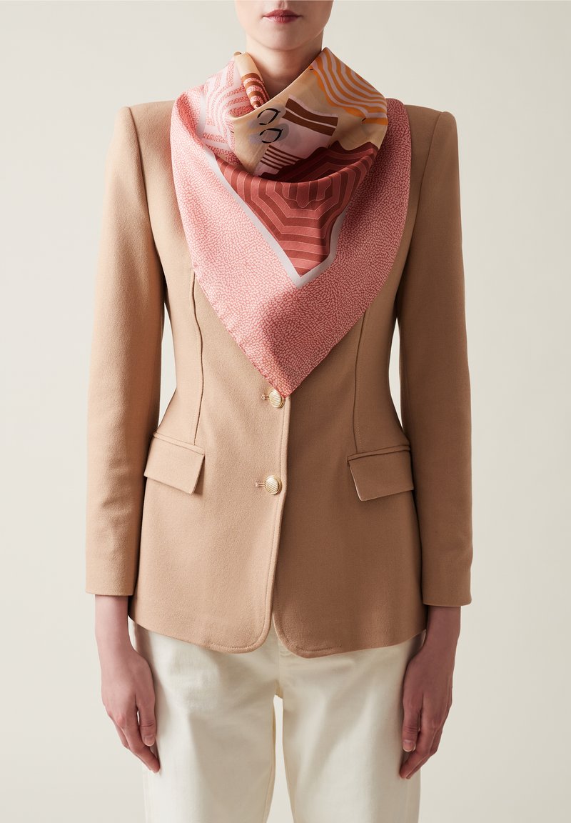 Foulard en soie dans des tons roses et oranges, arborant des motifs géométriques. Porté avec un blazer beige, mettant en valeur une texture lisse et une forme triangulaire.