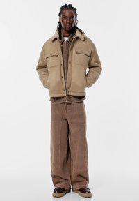 Jachetă izolată bej cu guler căptușit cu blană, două buzunare la piept. Pantaloni oversized maro cu cusături contrastante, asortați cu pantofi negri.