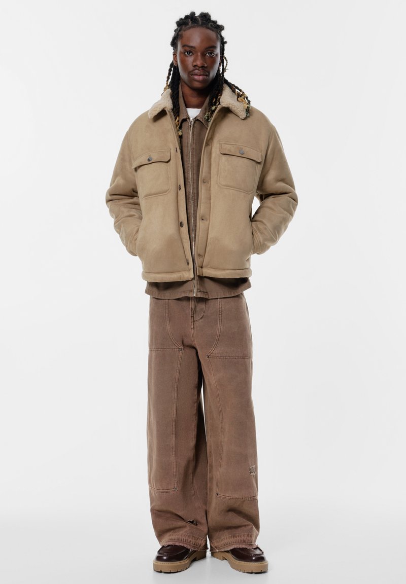 Jachetă izolată bej cu guler căptușit cu blană, două buzunare la piept. Pantaloni oversized maro cu cusături contrastante, asortați cu pantofi negri.