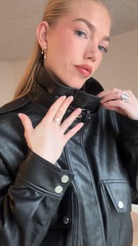 Črna usnjena jakna z visokim ovratnikom, stranskimi gumbi in sprednjimi žepekoma. Modelka prilagaja ovratnik, razkrivajoč gladko, teksturirano površino.