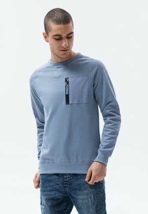Sweatshirt bleu clair avec un col rond et des manches longues. Il possède une poche zippée bleu foncé sur le côté gauche de la poitrine et des poignets côtelés.