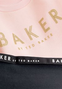 Rozā sporta krekls ar zelta strukturētu uzrakstu "BAKER" un melnu apakšējo paneli ar lentas detaļu, kurā rakstīts "BAKER by TED BAKER."