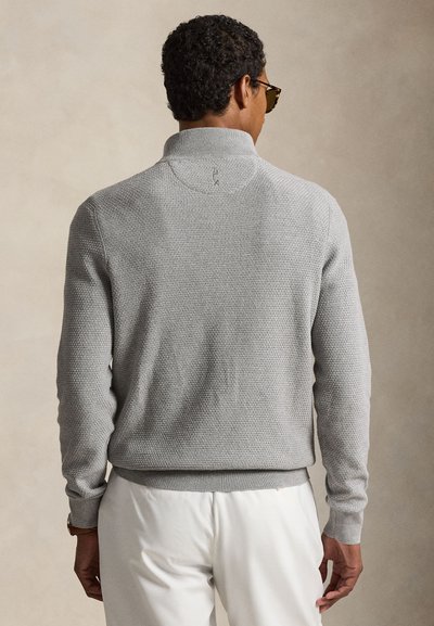 Uomo che indossa un maglione grigio chiaro strutturato e pantaloni bianchi, mostrato di spalle, su uno sfondo beige uniforme.