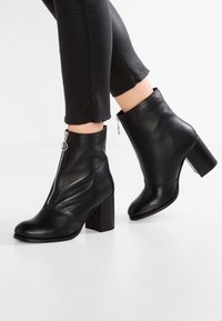 Černé kožené kotníkové boty s robustním podpatkem, bočním zipem a hladkým povrchem. Mají zaoblenou špičku a elegantní design.