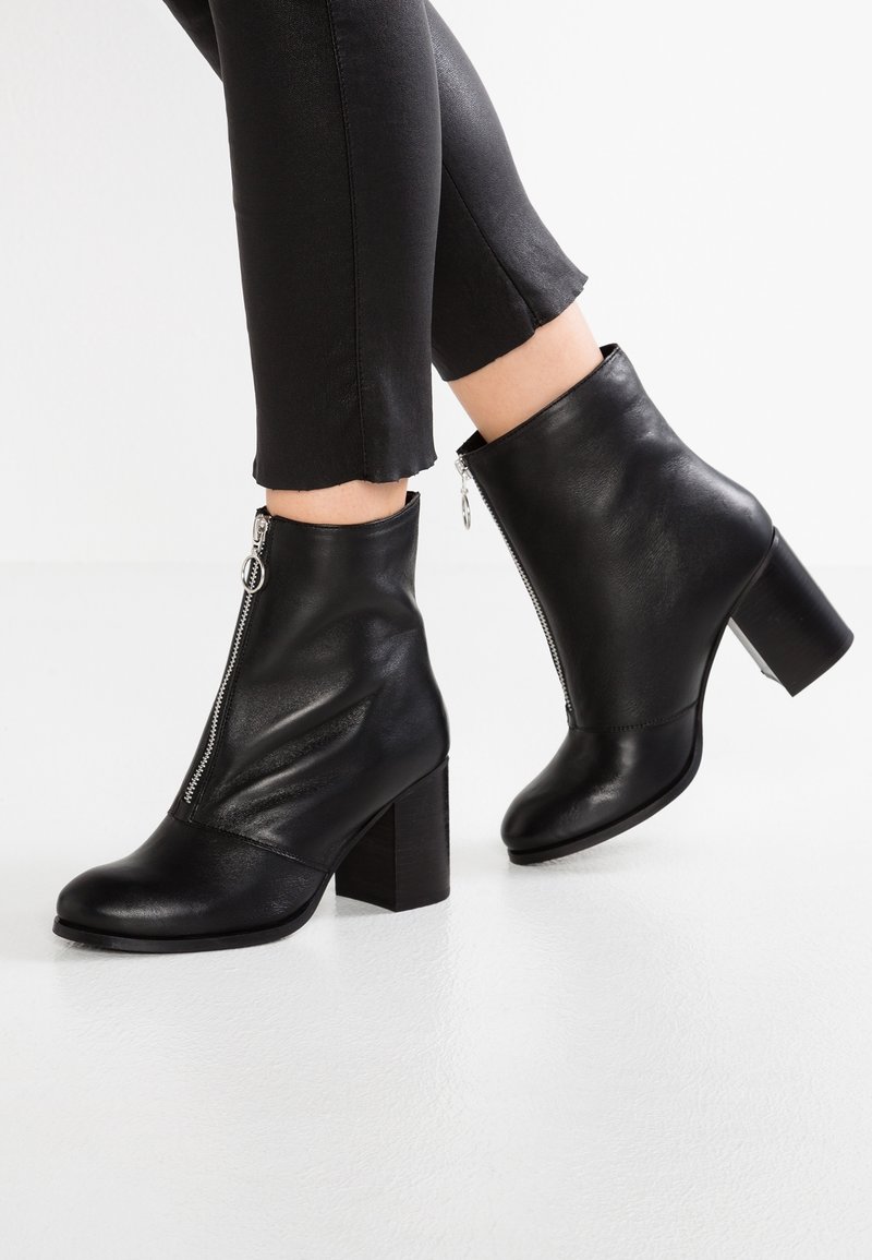 Černé kožené kotníkové boty s robustním podpatkem, bočním zipem a hladkým povrchem. Mají zaoblenou špičku a elegantní design.