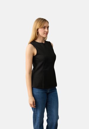 Femme aux longs cheveux blonds portant un haut sans manches noir et un jean bleu, debout devant un fond uni gris clair.